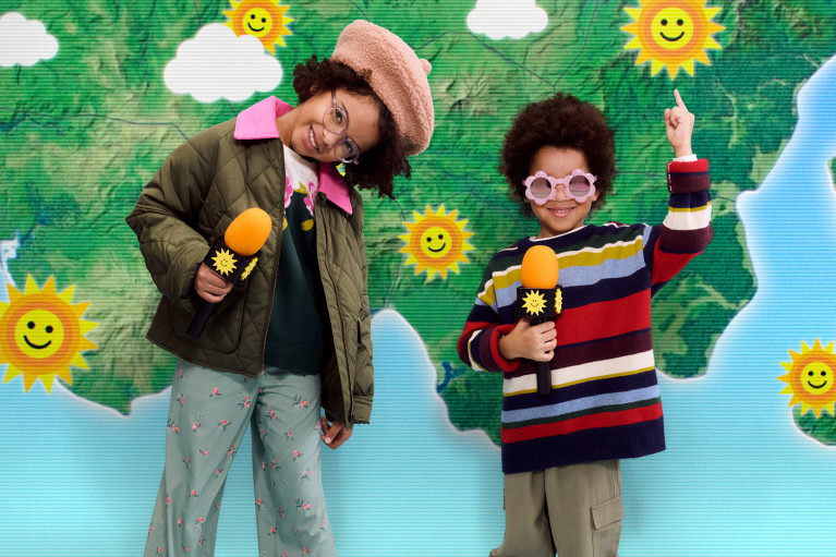 Two kids in playful clothes and microphones posing in front of a childrens' weather map showing only sunshine